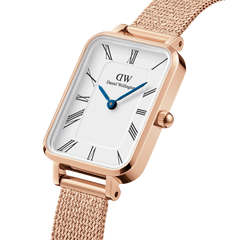 Close-up of the Quadro Roman Numerals Melrose watch showing its rose gold rectangular case, white dial with black Roman numerals, blue steel hands, and a fine mesh strap.