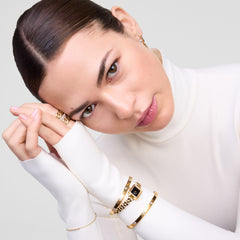 A person wearing the Classic Lumine Bracelet Gold 55mm on their wrist, styled with a gold watch and other gold bangles over a white long-sleeve top, showcasing the bracelet’s polished finish and sparkling cubic zirconia accents.