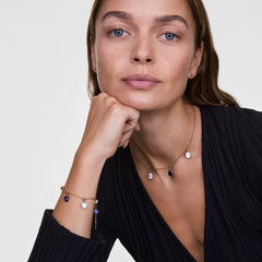 Woman wearing a gold chain bracelet and necklace, each adorned with the Blue Spot Heart Gold Charm and other semi-precious stone charms, styled with a black top for an elegant, layered jewellery look.