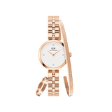 Rose gold women’s watch with a white dial and crystal hour markers, paired with a matching rose gold bracelet and ring, each featuring subtle crystal accents and Daniel Wellington branding.