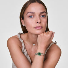 A woman wearing the Daniel Wellington Ophelia Mini Emerald Gold watch, featuring a gold link bracelet and an oval emerald green dial with gold Roman numerals, styled with a sleeveless top and a delicate necklace.
