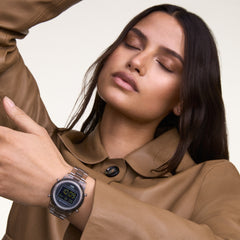 A woman wearing the Daniel Wellington Retro Digital Light Grey watch, featuring a transparent bio-based plastic strap and bold digital display, styled with a tan coat.