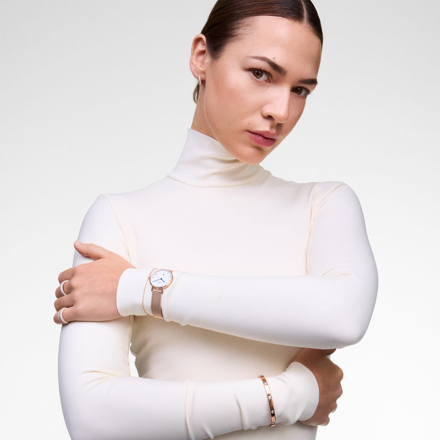 A person wearing the Mirelle Eternity Bracelet Rose Gold on their wrist, styled with a matching rose gold watch and bangle over a fitted cream turtleneck top, showcasing a refined and elegant layered jewellery look.