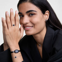 A woman wears the Daniel Wellington Quadro Mini & Audrey Black Set, featuring a rectangular black dial watch with a black mesh strap and a silver bracelet with a black circular charm, styled alongside matching black and silver jewellery for a coordinated, elegant look.