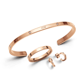 Rose gold jewellery set featuring a sleek open bracelet, matching ring, and small hoop earrings, each adorned with subtle Daniel Wellington branding and delicate crystal accents. The polished finish and coordinated design create an elegant, modern look.