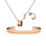 Rose gold jewellery set featuring a delicate chain necklace with a signature ring pendant, matching hoop earrings, and a sleek open cuff bracelet, each engraved with subtle Daniel Wellington branding. The coordinated pieces showcase a modern yet timeless design with a polished finish.