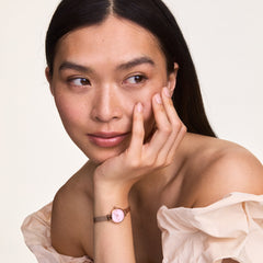 A woman wearing the Petite Mini Blush Melrose watch with a blush pink dial and rose gold mesh strap, styled with an off-the-shoulder top for an elegant, feminine look.