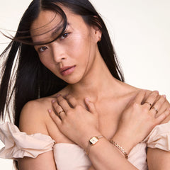 A woman wearing the Quadro Mini Melrose Champagne watch with a petite rectangular champagne dial and rose gold mesh strap, styled with matching rose gold jewellery and an off-shoulder cream dress.
