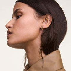 A woman wears a single Daniel Wellington 3-Link gold earring with a polished, geometric design, styled with a neutral collared top.