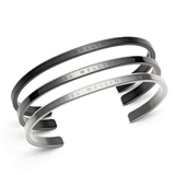 Set of three Daniel Wellington open cuff bracelets in black, graphite, and silver, each featuring a minimalist design and subtle engraved branding, stacked together to showcase their sleek, modern finish.