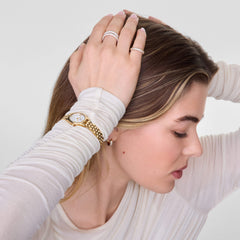 A woman wearing the Pavé Ring Gold 3 mm, styled on her hand alongside a gold watch and another pavé ring, showcases the ring’s slim gold band and sparkling white cubic zirconia crystals against a neutral long-sleeve top.
