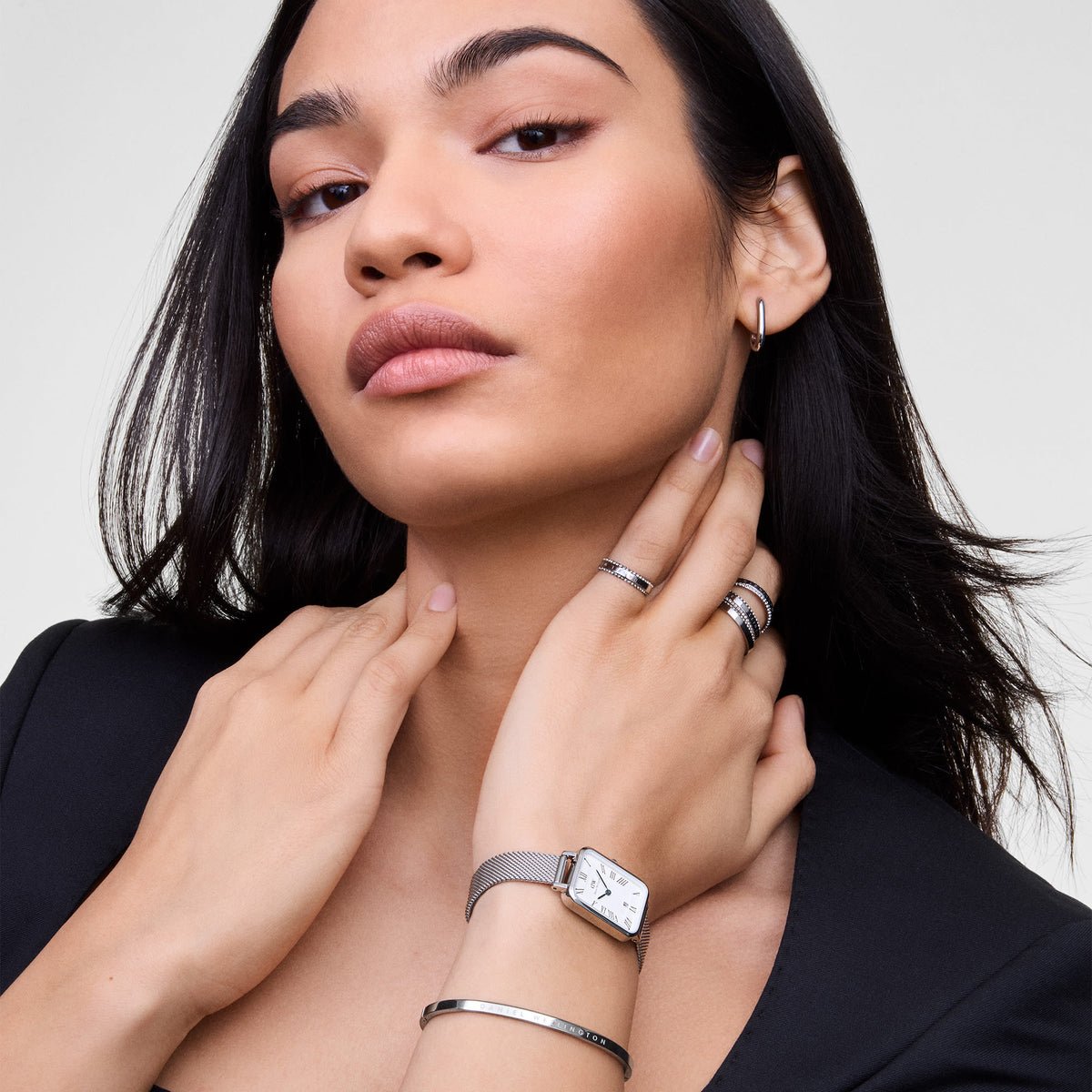 A woman wearing the Classic Aura Ring Silver by Daniel Wellington, styled with other silver jewellery including a watch, bangle, and hoop earring, showcasing the ring’s row of white cubic zirconia crystals and engraved band for a refined, modern look.