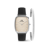 Square silver Daniel Wellington watch with a linen-textured beige dial and black leather strap, paired with a slim silver cuff bracelet engraved with the brand name. The set features minimalist styling and a refined, modern look.