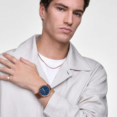 Man wearing the Classic Day Display St Mawes Rose Gold Arctic watch, featuring a blue patterned dial, rose gold case, and brown leather strap, styled with a light jacket and simple jewellery for a refined, modern look.