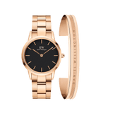 Rose gold stainless steel watch with a black dial and matching link strap, paired with a minimalist rose gold cuff bracelet engraved with "Daniel Wellington", both showcasing a modern and elegant design.