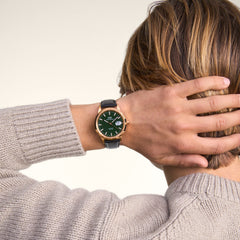 A man wearing the Daniel Wellington Iconic Sheffield Automatic Rose Gold watch with an emerald green dial, rose gold case, and black leather strap, styled with a beige knitted jumper.