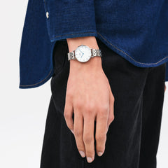 Woman wearing Petite Roman Numerals 5-Link Silver watch with white dial and blue hands, styled with denim shirt and black jeans.