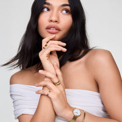 Woman wearing the Petite Roman Numerals Date 5-link Gold watch on her wrist, styled with matching gold jewellery and a white off-shoulder top, highlighting the watch’s elegant gold bracelet and classic white dial with Roman numerals.