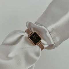 Quadro Mini Melrose Onyx women’s watch with a rectangular black dial and rose gold mesh strap, styled on a wrist with an elegant white blouse.