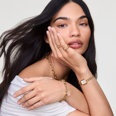 A woman wears the Mirelle Eternity Ring Gold 1.4 mm on her finger, styled with other gold jewellery including a chunky chain bracelet, a gold watch, and a matching necklace, highlighting the ring’s delicate band and sparkling cubic zirconia stones in a modern, layered look.