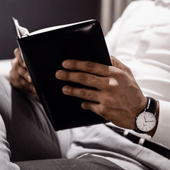 Man wearing Classic Sheffield Silver watch with black leather strap and white dial, reading a book in a relaxed setting.