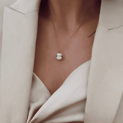 A woman wears the Aspiration Necklace Rose Gold Satin White, featuring a satin white ceramic sphere pendant with a rose gold-plated band on a delicate rose gold chain, styled with an elegant cream blazer.