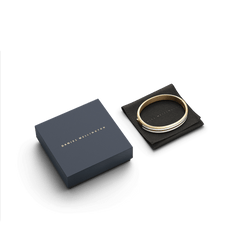 Emalie Infinite Gold Satin White bangle displayed on a black leather pouch next to an elegant navy Daniel Wellington gift box.
