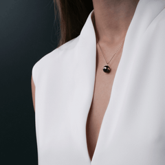 A woman wears the Aspiration Necklace Rose Gold Black, featuring a black ceramic sphere pendant with a rose gold plated band on a fine rose gold chain, styled with a white V-neck top.