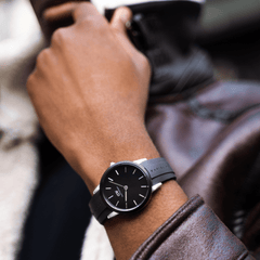 A person wearing the Daniel Wellington Iconic Motion watch with a black dial and black FKM rubber strap, styled with a brown leather jacket for a modern, casual look.
