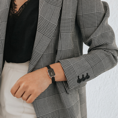 A woman wearing the Quadro Pressed Ashfield rectangular watch with a black dial, rose gold case, and black mesh strap, styled with a checked blazer and black lace top for a sophisticated, modern look.