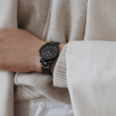 Black ceramic Daniel Wellington Iconic Link watch with a polished link bracelet and minimalist black dial, worn on the wrist with a cream knit jumper.